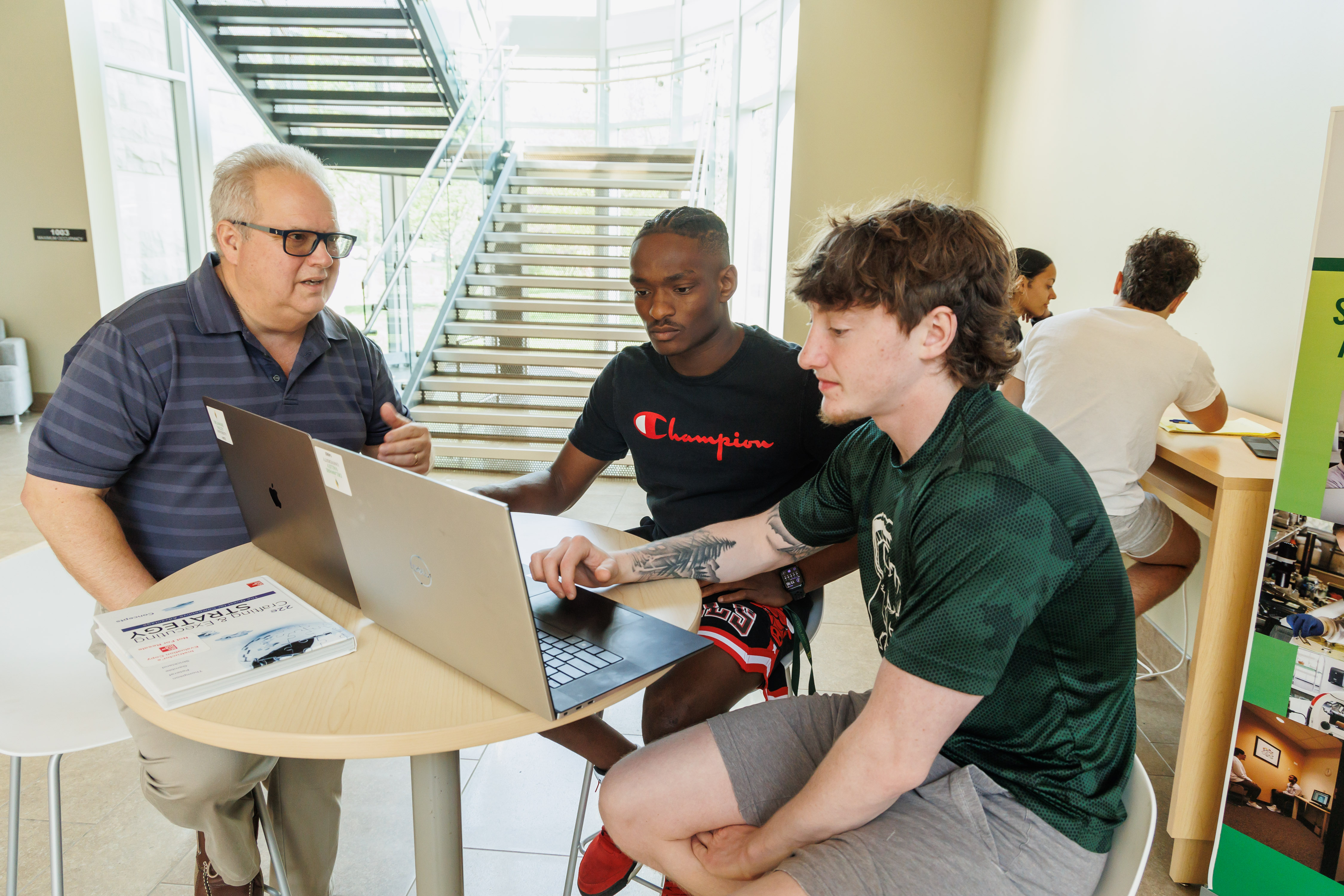 A business administration professor is working with two male students on a laptop in the life sciences building lobby.