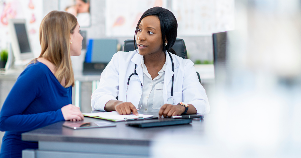 A female doctor is speaking with a female patient at a table while pointing to papers.