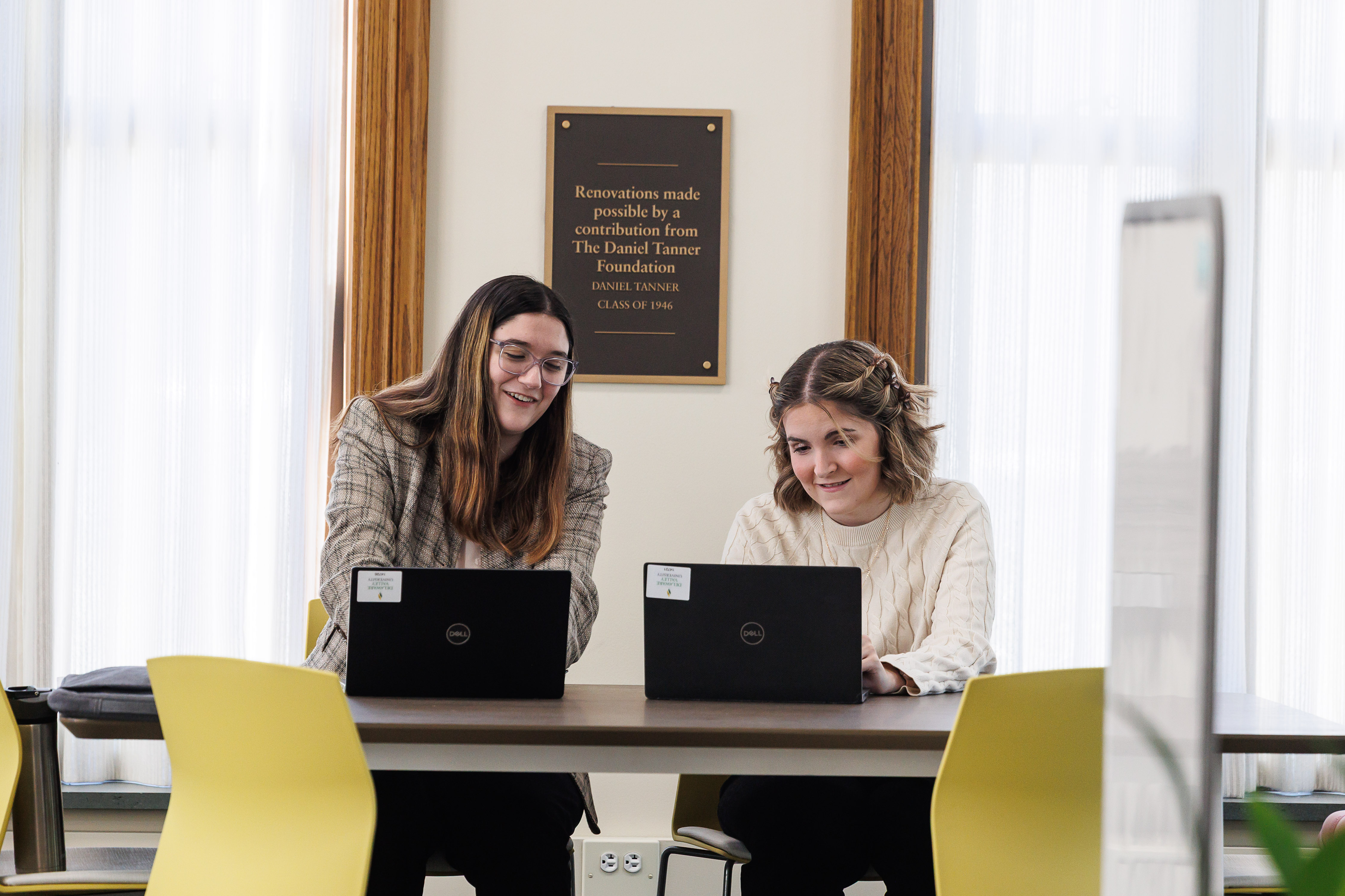 Two women are in the library on their laptops working on a project together.