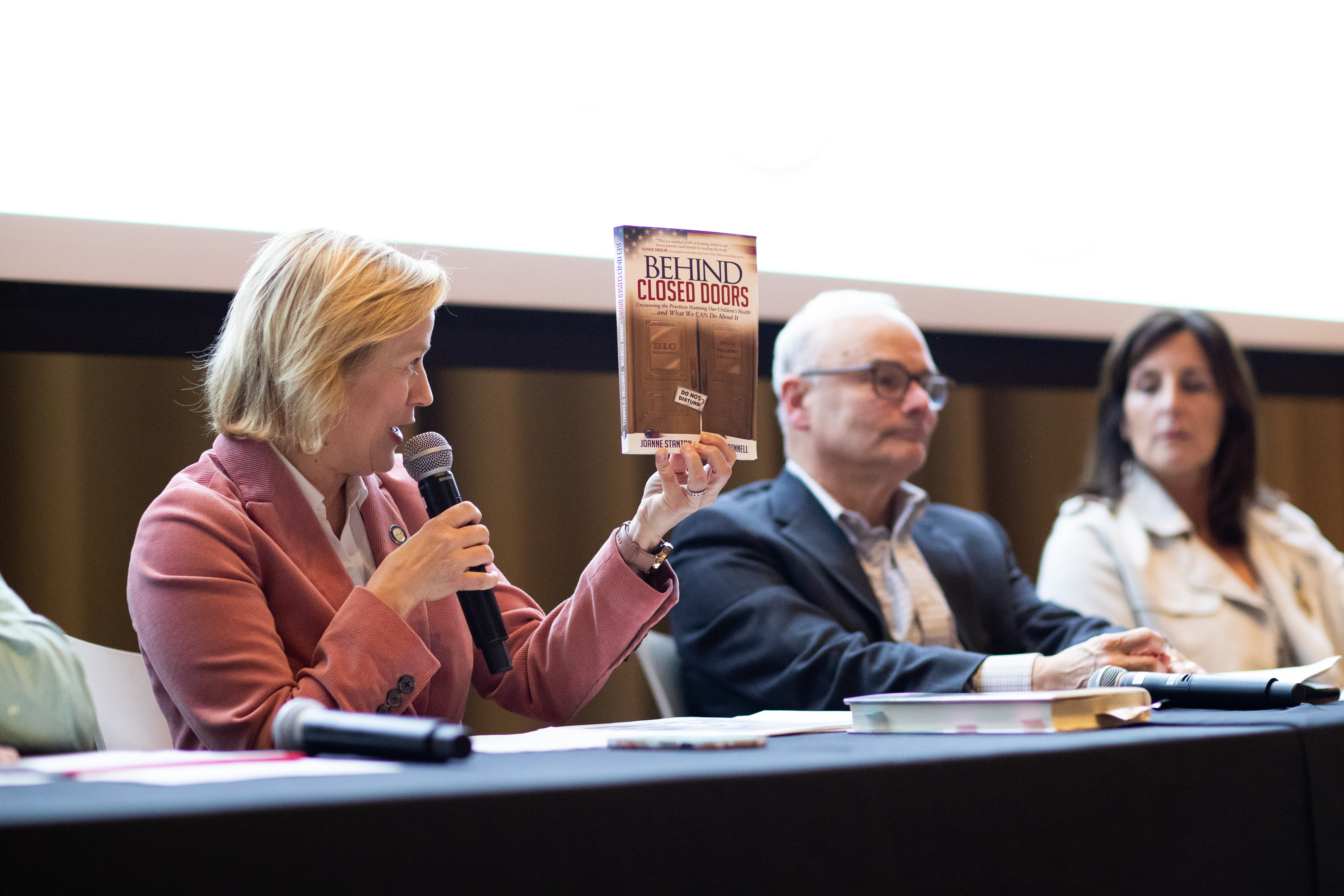 Woman in pink blazer sitting at table holding up a book while speaking into a handheld microphone