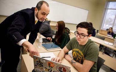 Man in suit leaning over desk speaking to stuent looking at computer.