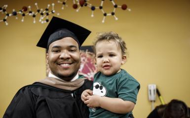 A graduate from the Continuing Studies department in his cap and gown, holding his son.
