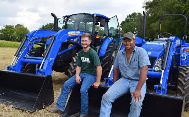 Two students in a field sitting on a tractor bucket.