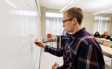 A professor is writing on the white board in front of a class. 