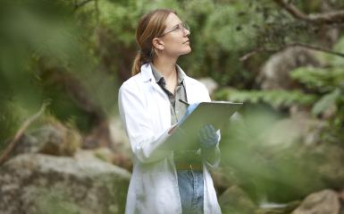 A woman is in a lab coat holding a clipboard outdoors. 