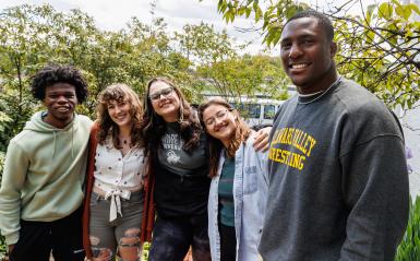 Five students, male and female, are smiling and laughing outdoors surrounded by greenery.