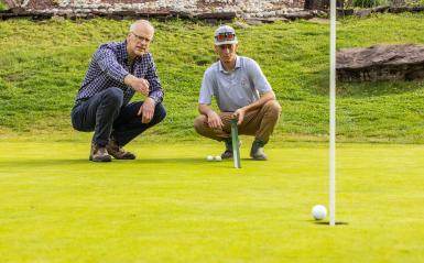 Professor Doug Linde and a Student are on a golf course speaking one-on-one while squatted down on the turf.