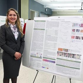Diana Rachii, who is wearing a grey suit, stands in front of a scientific research poster. The poster is about her research on fruit pigments. 