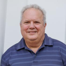 A headshot of lance hill in a blue collared shirt in front of the feldman building. 