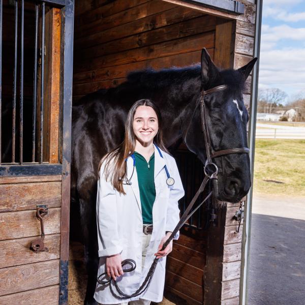 Nina Dellarte with a white coat and a horse in the stables of DelVal.