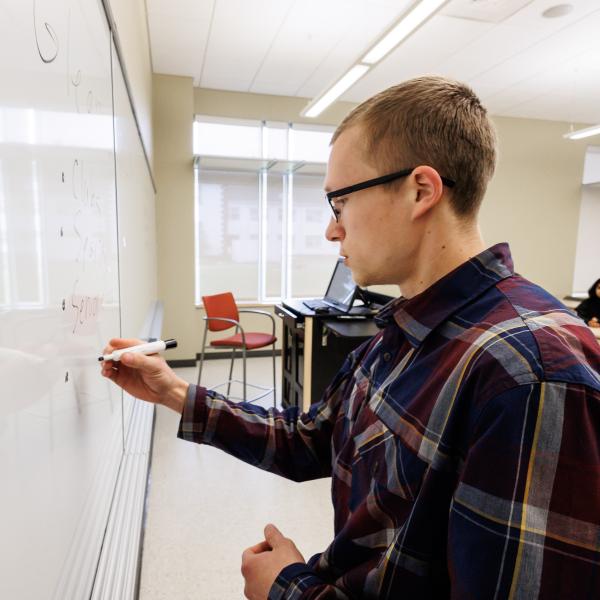 A male teacher is at the front of the class, writing on the whiteboard with dry erase marker. 