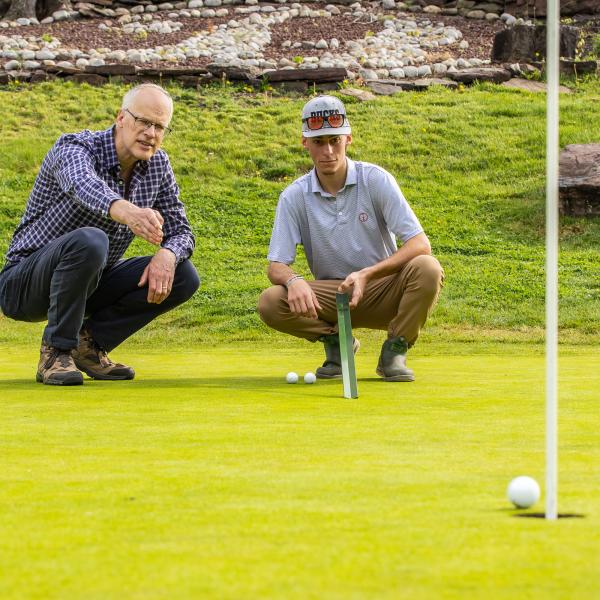 Professor Doug Linde and a Student are on a golf course speaking one-on-one while squatted down on the turf.