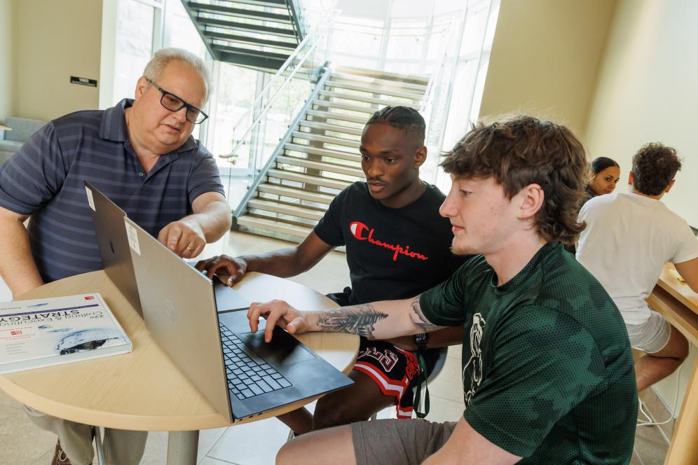 Lance Hill talking with two students infront of a computer in the Life Sciences Building. 