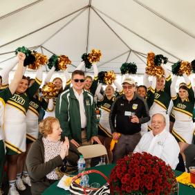Cheerleaders are cheering behind alumni sitting at a round table. 
