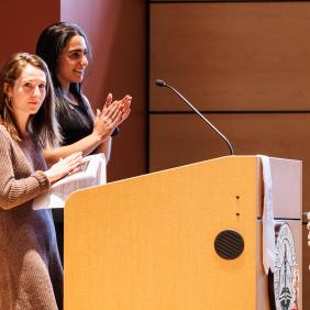 Two women are on stage at a podium clapping during an event. 