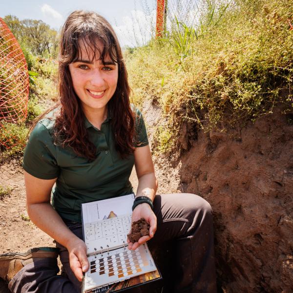 Dr. Jaclyn Fiola in a soil pit with a shovel, smiling at the camera. 