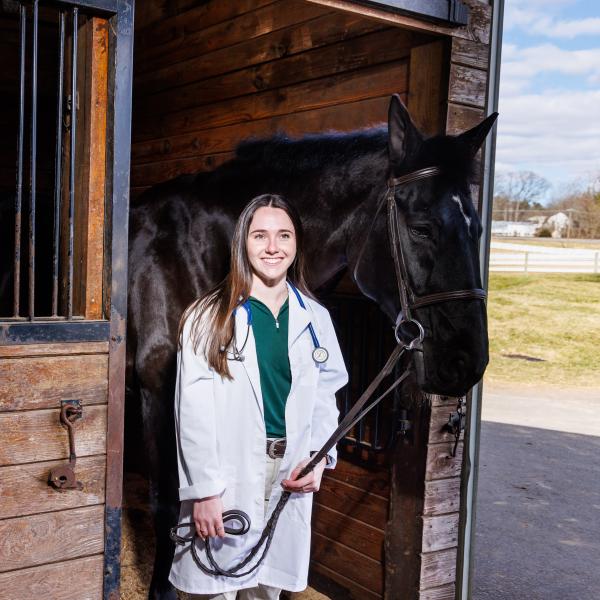 Nina Dellarte with a white coat and a horse in the stables of DelVal.