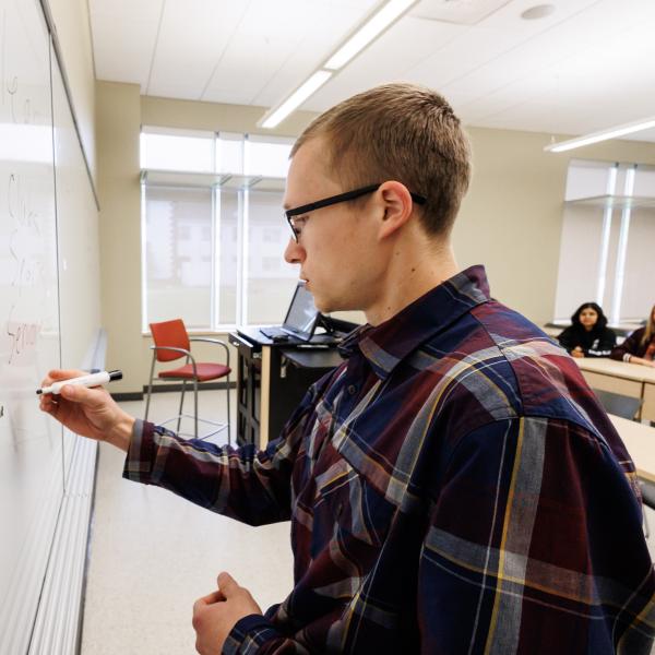 A male professor is writing on a white board in a classroom. 