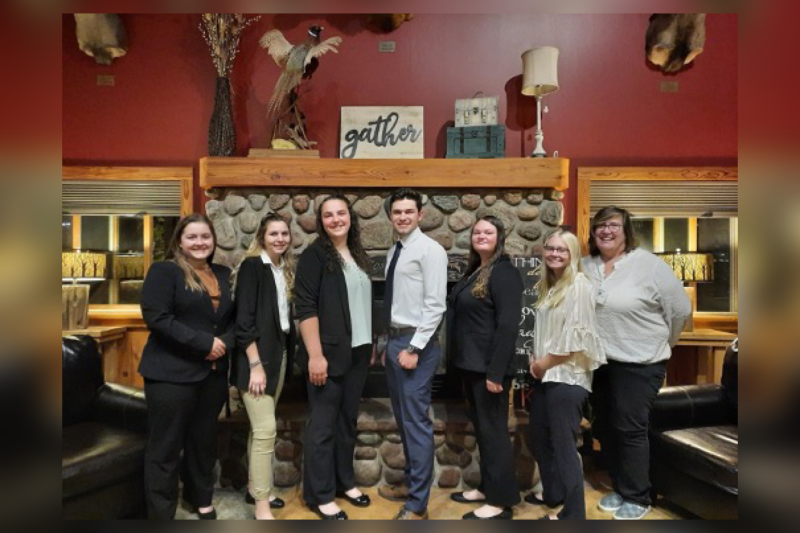 Six Dairy Science students dressed business casual and Ms. Powell line up for a photo in front of a fireplace. 
