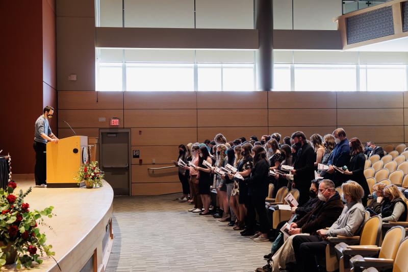 Students stand in front of their seats in the auditorium waiting to be called to receive their certificate. 