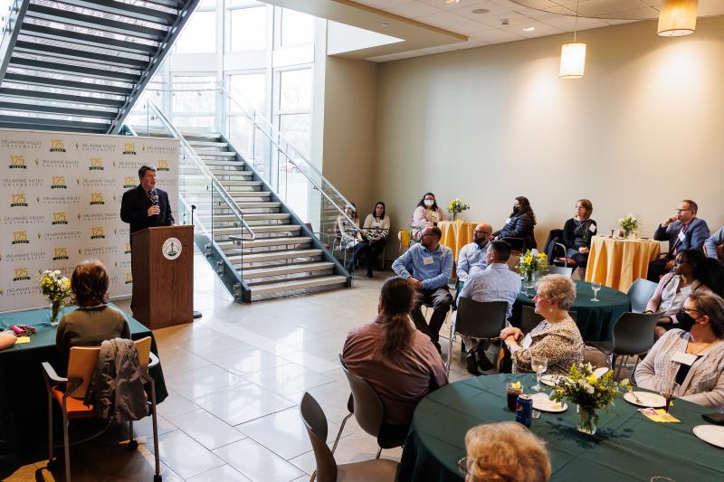Dr. Benjamin Rusiloski is holding a microphone while addressing a crowd of people seated at round tables. 