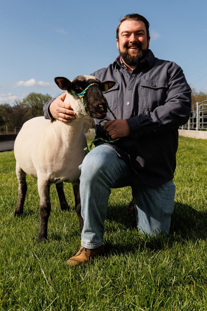 Bill Fritz kneels on the grass with a sheep under his right arm. 