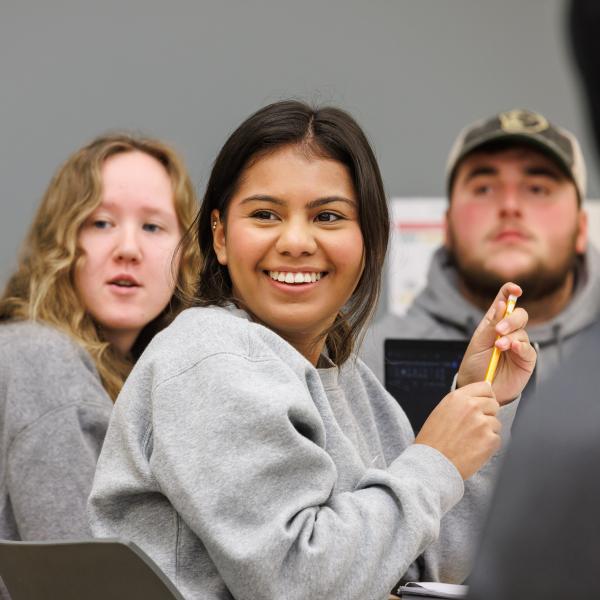 A student is in class smiling at the professor with a person in her hand. 